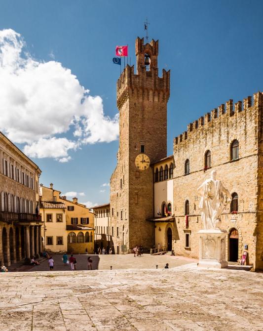 Square with medieval tower and statue under a blue sky.
